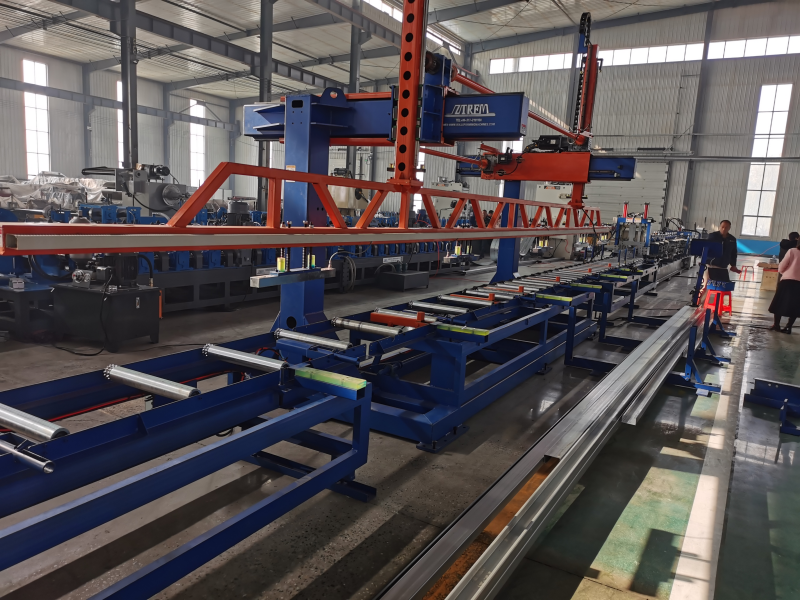A PLC adjustment CZ purlin making machine displayed in a large industrial hall, featuring an overhead gantry structure, roller conveyors, and finished metal profiles on output tables, with personnel observing nearby.